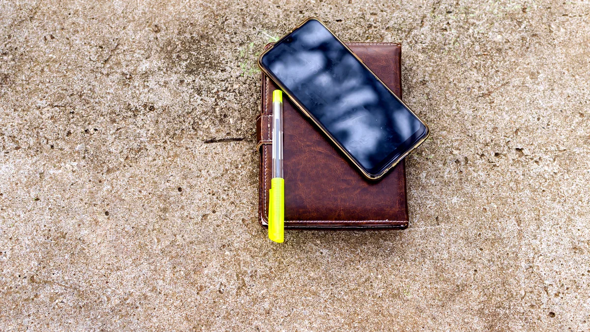 A phone in a worn leather case, the stitching frayed at the corner, on a wooden table.