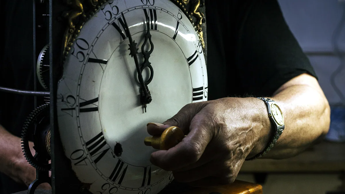 Hands turning the crown of a small mechanical clock, time set back and running again.