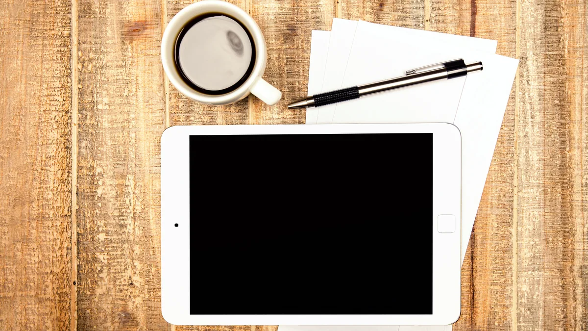 A small touchscreen tablet resting on a worn wooden desk, lit by a single warm lamp.