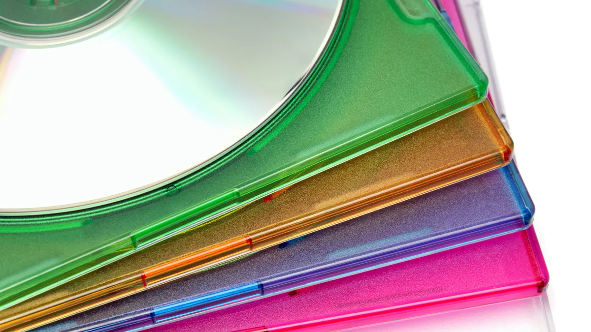 A stack of old CD jewel cases with faded labels, leaning against a bookshelf.